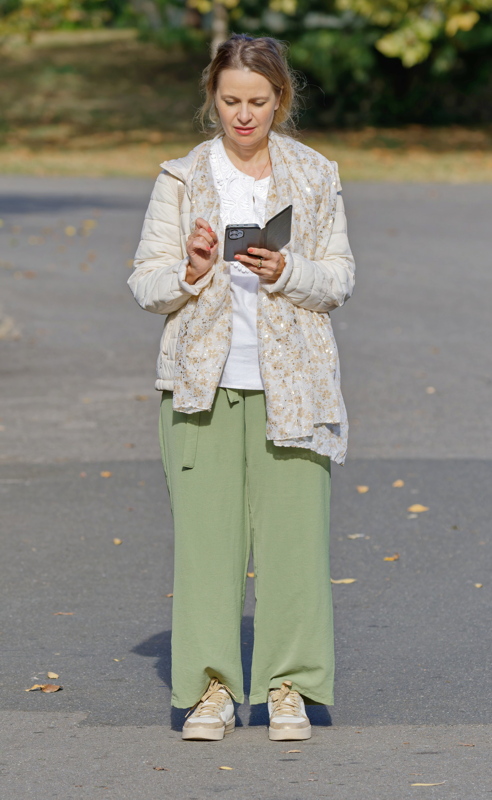 25 - A woman wearing a casual layered outfit featuring a light quilted jacket, a patterned scarf, a white top, loose green wide-leg trousers, and light-colored sneakers while looking at her phone outdoors